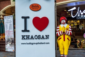 A large sign with 'I love Khaosan' next to a statue of a clown in a praying pose, located outside a shop. The clown is wearing a red and white striped outfit with yellow overalls featuring a fast food chain logo. A banner on the left advertises fitness tickets with pricing in Thai baht.