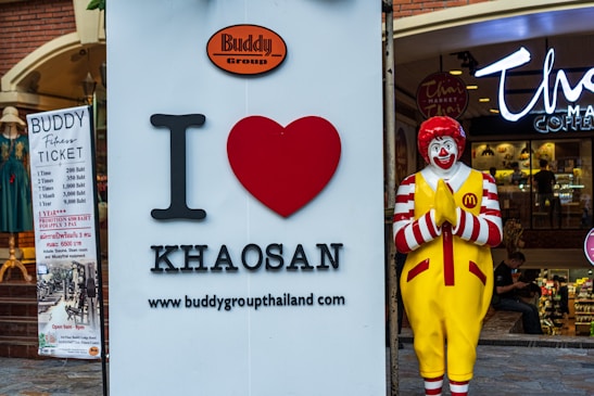 A large sign with 'I love Khaosan' next to a statue of a clown in a praying pose, located outside a shop. The clown is wearing a red and white striped outfit with yellow overalls featuring a fast food chain logo. A banner on the left advertises fitness tickets with pricing in Thai baht.
