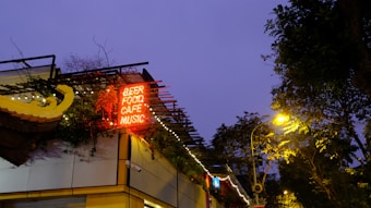 A building facade with a neon sign displaying the words 'BEER, FOOD, CAFE, MUSIC' in red. The structure is adorned with decorative elements and small lights. Trees and streetlamps are visible, with a deep blue sky in the background.