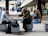 Two men are engaged in a game of chess at a concrete table in a public space. One man, wearing a dark jacket and cap, is concentrated on the board while the other, in a gray jacket, observes intently. Nearby, another person is sitting, focused on their own chess game, with a chessboard bag and a bright blue shopping bag beside them. People walk by in the background amid a modern urban setting with concrete structures and banners hanging from the building.