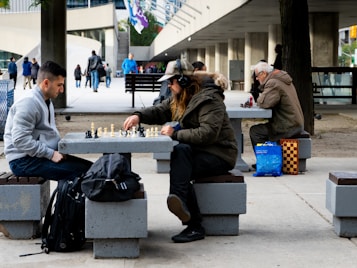 Two men are engaged in a game of chess at a concrete table in a public space. One man, wearing a dark jacket and cap, is concentrated on the board while the other, in a gray jacket, observes intently. Nearby, another person is sitting, focused on their own chess game, with a chessboard bag and a bright blue shopping bag beside them. People walk by in the background amid a modern urban setting with concrete structures and banners hanging from the building.