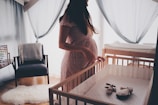 A pregnant woman stands beside a wooden crib in a softly lit room. The crib contains two stuffed toys, and the room features soft blue curtains, an armchair with a draped blanket, and a small table with an abacus. Light filters gently through the window, creating a calm and serene atmosphere.