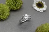 A silver ring with a large round diamond at its center is displayed on a gray textured surface, surrounded by several green and white flowers.