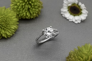 A silver ring with a large round diamond at its center is displayed on a gray textured surface, surrounded by several green and white flowers.