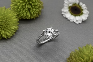 A silver ring with a large round diamond at its center is displayed on a gray textured surface, surrounded by several green and white flowers.