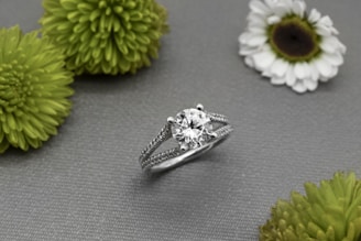 A silver ring with a large round diamond at its center is displayed on a gray textured surface, surrounded by several green and white flowers.
