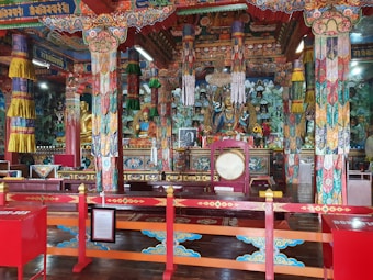 Intricately decorated interior of a Buddhist temple featuring colorful columns, ornamental patterns, and religious iconography. Statues of deities are prominently displayed, surrounded by vibrant textiles and offerings on the altar. The space is adorned with detailed artwork, demonstrating a rich cultural aesthetic.