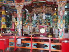Intricately decorated interior of a Buddhist temple featuring colorful columns, ornamental patterns, and religious iconography. Statues of deities are prominently displayed, surrounded by vibrant textiles and offerings on the altar. The space is adorned with detailed artwork, demonstrating a rich cultural aesthetic.