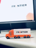 A small toy truck in orange and white with the word 'FRONTIER' printed on its side sits on an office desk. Behind it, a large computer monitor displays the same logo with a modern design.