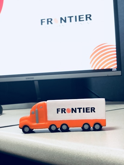 A small toy truck in orange and white with the word 'FRONTIER' printed on its side sits on an office desk. Behind it, a large computer monitor displays the same logo with a modern design.