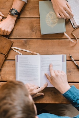 A group of people sitting at a wooden table with open books and notebooks. One person is pointing at a page in a book, while another person's hands, wearing a watch, rest on the table. Pens and a leather bag are also visible on the table, suggesting a study or discussion setting.