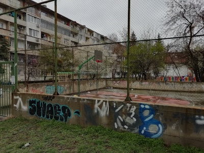 An outdoor basketball court surrounded by a chain-link fence and graffiti-covered walls. The court shows signs of wear and is wet, possibly after rain. In the background, there are apartment buildings with balconies and trees with sparse foliage, suggesting it might be autumn or winter.