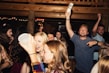 A lively group of young people at a party, some holding drinks and chatting animatedly. The atmosphere is vibrant with dim lighting and wooden interior decor. The crowd appears to be enjoying the music and having a good time.