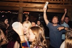A lively group of young people at a party, some holding drinks and chatting animatedly. The atmosphere is vibrant with dim lighting and wooden interior decor. The crowd appears to be enjoying the music and having a good time.