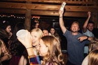 A lively group of young people at a party, some holding drinks and chatting animatedly. The atmosphere is vibrant with dim lighting and wooden interior decor. The crowd appears to be enjoying the music and having a good time.