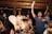 A lively group of young people at a party, some holding drinks and chatting animatedly. The atmosphere is vibrant with dim lighting and wooden interior decor. The crowd appears to be enjoying the music and having a good time.