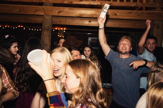 A lively group of young people at a party, some holding drinks and chatting animatedly. The atmosphere is vibrant with dim lighting and wooden interior decor. The crowd appears to be enjoying the music and having a good time.