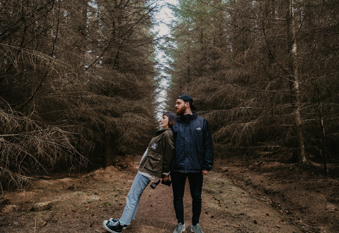 woman in black jacket and blue denim jeans standing on brown dirt road between trees during