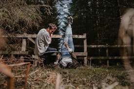 Two people are outdoors near a wooden fence. One person is sitting on the ground, laughing, while the other is playfully leaning against the fence with legs up in the air. The area is surrounded by trees, suggesting a forest setting. The ground is covered with grass and dry leaves.