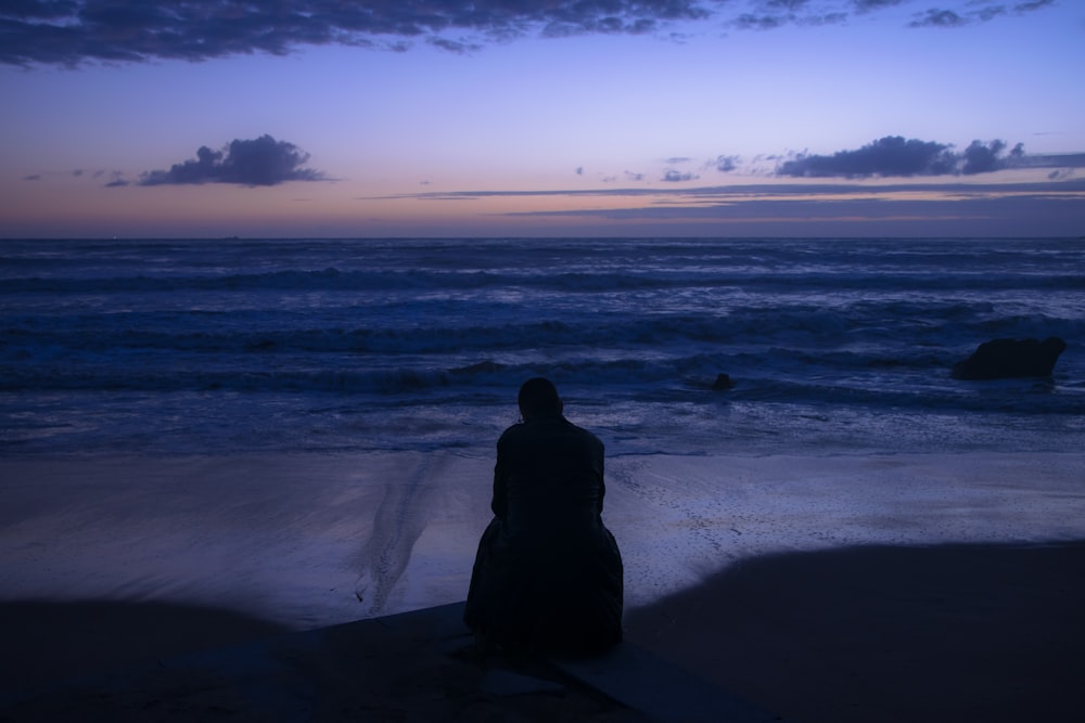 silhouette of person sitting on seashore during sunset