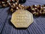 An intricately designed octagonal gold pendant featuring Sanskrit inscriptions is placed on a string of beads. The beads are a mix of round brown wooden beads and golden beads with detailed crafting.