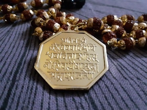An intricately designed octagonal gold pendant featuring Sanskrit inscriptions is placed on a string of beads. The beads are a mix of round brown wooden beads and golden beads with detailed crafting.
