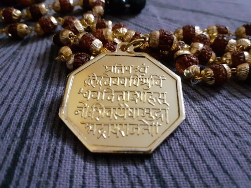 An intricately designed octagonal gold pendant featuring Sanskrit inscriptions is placed on a string of beads. The beads are a mix of round brown wooden beads and golden beads with detailed crafting.