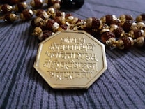 An intricately designed octagonal gold pendant featuring Sanskrit inscriptions is placed on a string of beads. The beads are a mix of round brown wooden beads and golden beads with detailed crafting.