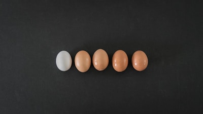 A row of five eggs is aligned horizontally on a dark surface. The first egg on the left is white, while the remaining four eggs are brown. They are evenly spaced and centered in the image.