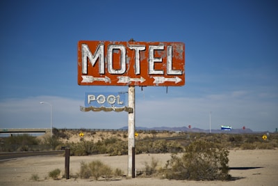A weathered and rusted vintage motel sign stands prominently in a desert landscape. The red sign reads 'MOTEL' with large white letters and has an arrow pointing to the right. Below it, another sign reads 'POOL' with an arrow. The background is a flat, arid environment with scattered shrubs and distant mountains under a clear blue sky.