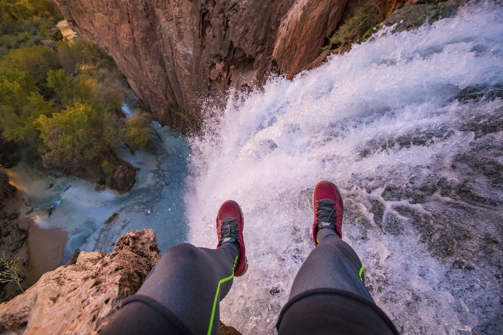 Persona con pantalones negros y zapatillas rojas sentada en una roca cerca de las cascadas de agua durante el día
