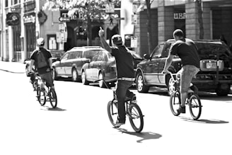 Three individuals ride bicycles down a city street. The scene appears to be lively with people enjoying a sunny day. The riders wear casual clothing and one of them has a backpack. Buildings and parked cars line the street, creating an urban backdrop.