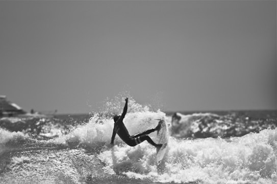 A surfer skillfully maneuvers a wave, with their body angled sharply and one arm extended outward. The ocean's surf is lively, creating a backdrop of splashing water and waves. Bright sunlight highlights the sea's texture, casting a dramatic silhouette of the surfer.