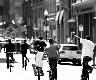 A group of people are riding bicycles along a busy city street. The scene is lively and dynamic, with a mix of pedestrians and vehicles visible in the background. The black and white tones give the image a timeless, nostalgic feel.