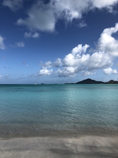 A tranquil beach scene with clear turquoise water gently meeting the sandy shore. In the distance, a sailboat is visible on the horizon near a range of lush green hills under a sky filled with fluffy white clouds.