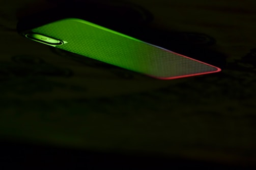 A mobile phone partially illuminated with green and red lighting, creating a contrast against the dark background. The illumination highlights the phone's texture and the outline of the camera.