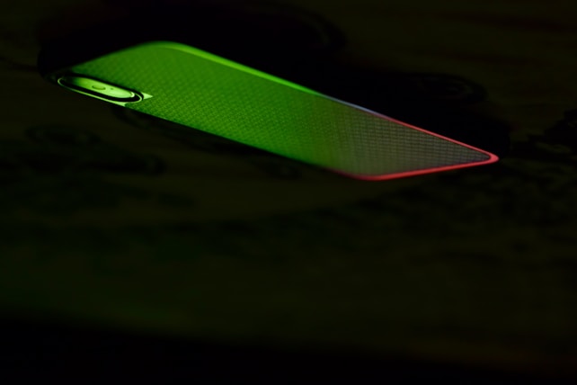 A mobile phone partially illuminated with green and red lighting, creating a contrast against the dark background. The illumination highlights the phone's texture and the outline of the camera.