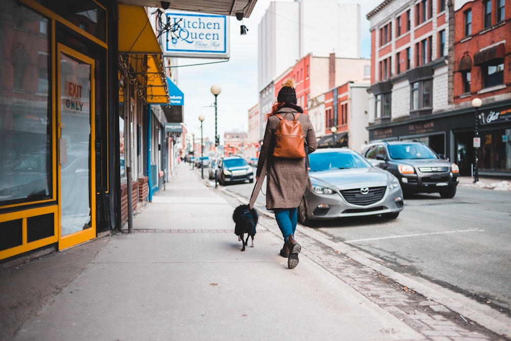 woman in brown coat walking on sidewalk with dog during daytime