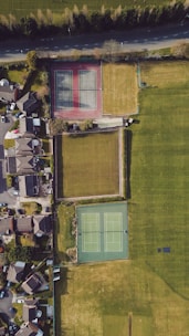 An aerial view of a residential neighborhood featuring two tennis courts, surrounded by grassy areas and adjacent to a row of houses. The tennis courts are positioned side by side, one with a red surface and the other with a green surface. A road runs parallel to the top edge, bordered by trees.