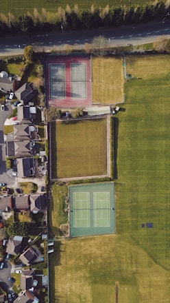 An aerial view of a residential neighborhood featuring two tennis courts, surrounded by grassy areas and adjacent to a row of houses. The tennis courts are positioned side by side, one with a red surface and the other with a green surface. A road runs parallel to the top edge, bordered by trees.