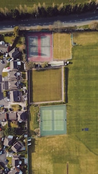 An aerial view of a residential neighborhood featuring two tennis courts, surrounded by grassy areas and adjacent to a row of houses. The tennis courts are positioned side by side, one with a red surface and the other with a green surface. A road runs parallel to the top edge, bordered by trees.