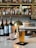A bar setting featuring a glass of beer, a glass of water, and several bottles of wine on a marble countertop. A brass lamp provides lighting, with shelves of assorted bottles in the blurred background.