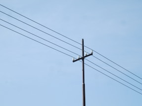 A utility pole with multiple power lines extending across a clear blue sky. The pole is made of wood and has several insulators attached at the top.