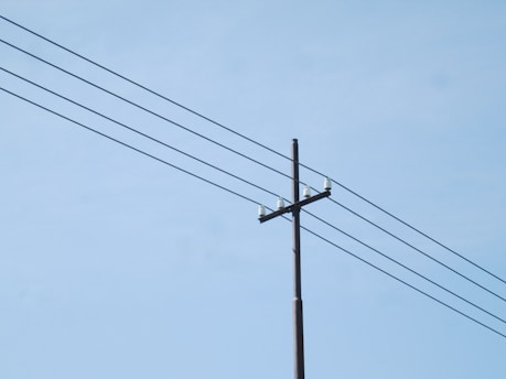 A utility pole with multiple power lines extending across a clear blue sky. The pole is made of wood and has several insulators attached at the top.
