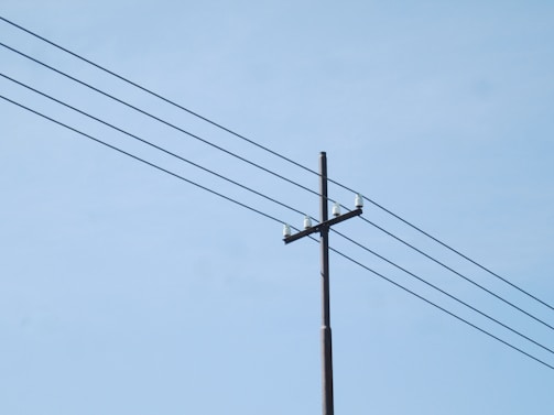 A utility pole with multiple power lines extending across a clear blue sky. The pole is made of wood and has several insulators attached at the top.