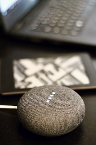 A small, round, gray smart speaker with illuminated dots is placed on a dark surface. In the background, a partially blurred laptop and a tablet with a geometric pattern on its case are visible.