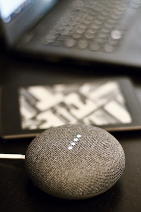 A small, round, gray smart speaker with illuminated dots is placed on a dark surface. In the background, a partially blurred laptop and a tablet with a geometric pattern on its case are visible.