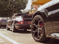 A scene of three sports cars parked along a street, with a lush green tree in the background. The focus is on the sleek design of the cars, particularly highlighting their wheels and body. People are seen in the background, adding a lively urban feel to the scene.
