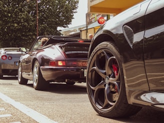 A scene of three sports cars parked along a street, with a lush green tree in the background. The focus is on the sleek design of the cars, particularly highlighting their wheels and body. People are seen in the background, adding a lively urban feel to the scene.