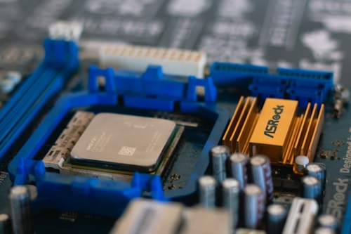 A close-up view of a computer motherboard featuring an AMD processor and a prominent ASRock heatsink. Blue slots, possibly for RAM, are visible alongside various capacitors and other electronic components.